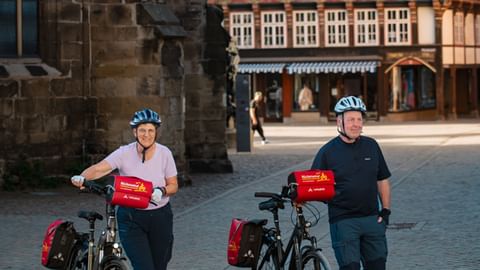 Zwei Radfahrer mit Helmen und roten Packtaschen stehen mit ihren Rädern auf dem Kopfsteinpflasterplatz in Hameln vor mittelalterlichem Turm und Fachwerkhäusern.