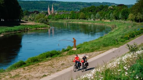 Zwei Radfahrer auf befestigtem Weg entlang der Weser mit Angler am Flussufer. Historisches Höxter mit Doppeltürmen im Hintergrund.