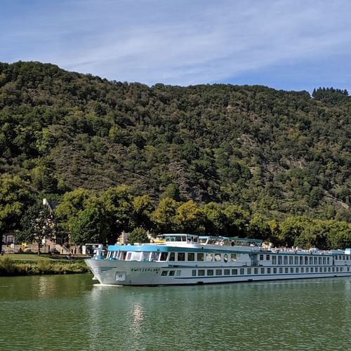 MS Switzerland Flusskreuzfahrtschiff auf ruhigem Wasser mit bewaldeten Bergen und kleinem Dorf im Hintergrund unter blauem Himmel.