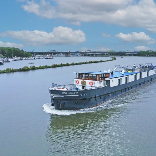 MS Magnifique IV, a white and blue river cruise ship, sailing on a wide river with marina and green landscape in the background.