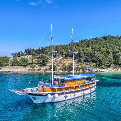 Weiße Segelyacht San Snova mit zwei Masten vor Anker in kristallklarem türkisfarbenem Wasser nahe einer bewaldeten Küste unter blauem Himmel.