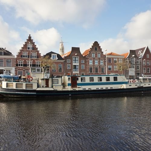 MS Flora, a white and green passenger vessel, moored along a canal with traditional Dutch gabled houses and a church tower in the background.