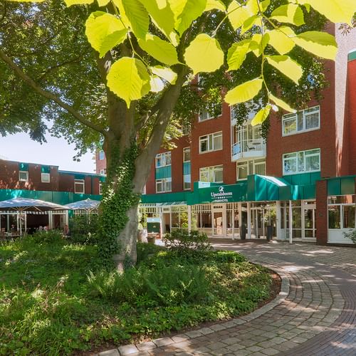 Außenansicht des Upstalsboom Hotels Emden mit roter Backsteinfassade und grünen Akzenten. Gepflasterter Innenhof mit alten Bäumen und Terrasse.