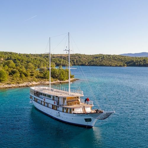 Luftaufnahme der weißen Segelyacht Magellan vor Anker im türkisfarbenen Wasser einer bewaldeten kroatischen Bucht mit Bergen im Hintergrund.
