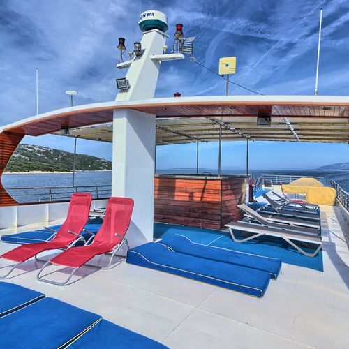 Sun deck of MY Andela Lora with blue and red loungers under a wooden canopy. Croatian coastline visible in the background.