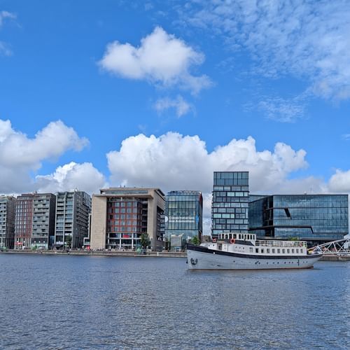 Weißes Passagierschiff Floor van Aemstel am modernen Ufer mit zeitgenössischen Glas- und Betongebäuden unter blauem bewölktem Himmel.