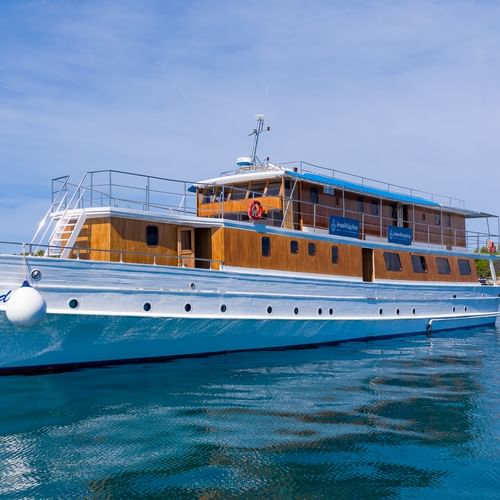 Weiße Motoryacht Linda mit Holzaufbau und blauen Akzenten vor Anker in kristallklarem blauen Wasser unter teilweise bewölktem Himmel.