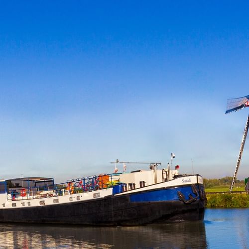 Blue and white cargo barge MS Sarah moored on a canal beside a traditional Dutch windmill with green body and brown base under clear sky.