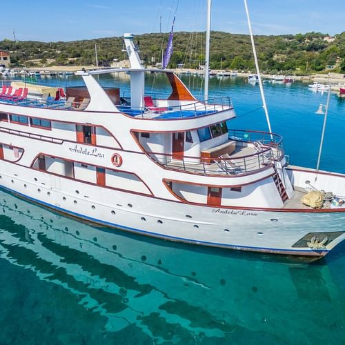 Weiße Motoryacht Andela Lora liegt in kristallklarem türkisem Wasser eines kroatischen Hafens mit Hügeln und Segelbooten im Hintergrund.