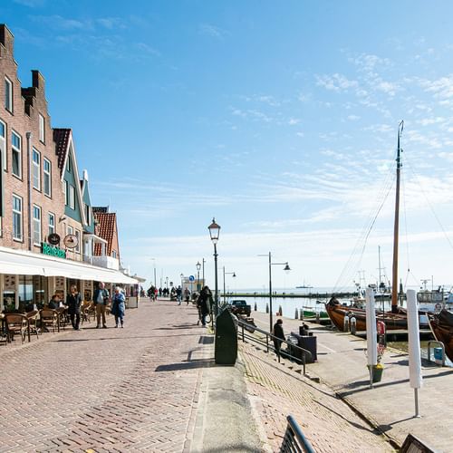 Uferpromenade in Volendam mit traditionellen holländischen Backsteinhäusern, Café-Terrassen und Holzbooten im Hafen unter blauem Himmel.