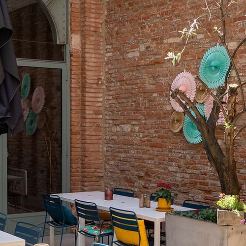 Außenterrasse mit weißem Tisch, bunten Stühlen und dekorativen Papierfächern an einem Baum vor einer Backsteinmauer in Toulouse.