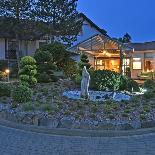 Landhotel Schnuck in der Dämmerung mit beleuchtetem Eingang und moderner Glasfassade. Runder Garten mit Skulptur und Zierpflanzen im Vordergrund.