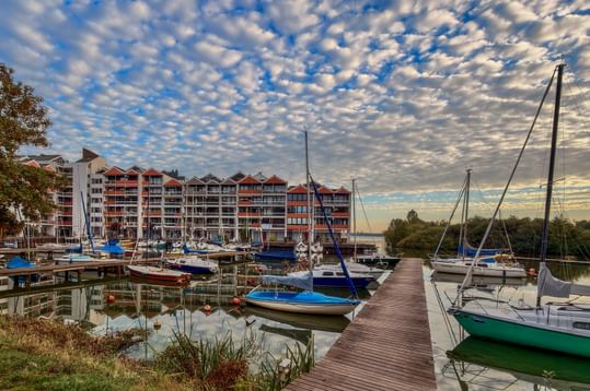 Hafen in Rastede mit Segelbooten am Holzsteg. Moderne Wohngebäude säumen das Ufer unter dramatischem Wolkenhimmel bei Sonnenuntergang.