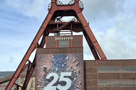 Roter Förderturm der Zeche Zollverein mit Backsteingebäude und '25'-Banner. Die ikonische Industriestruktur erhebt sich vor blauem Himmel.