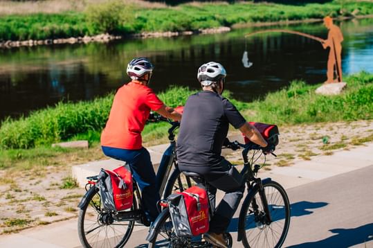 Zwei Radfahrer mit Helmen und Tourenrädern mit roten Packtaschen sitzen auf Kopfsteinpflaster an der Weser. Im Hintergrund angelt eine Person.