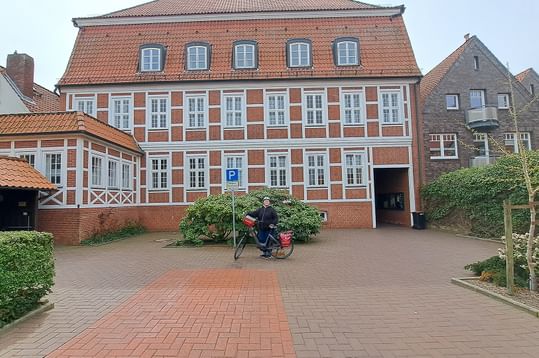 Frau namens Olga mit Fahrrad vor historischem rotem Backsteinrathaus in Bad Bederkesa mit weißen Fenstern und Ziegeldach.