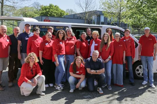 Großes Team-Gruppenfoto mit Personen in roten Shirts, die vor einem weißen Transporter stehen und knien, draußen an einem sonnigen Tag.