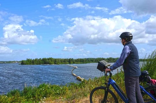 Radfahrer mit Helm steht neben blauem Fahrrad auf Grasweg mit Blick auf IJsselmeer mit Schilf und fernen Inseln unter bewölktem Himmel.