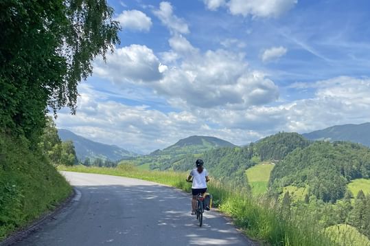 Radfahrer auf asphaltierter Straße entlang der Alpe Adria Route mit grünen Hügeln, Wäldern und Bergen unter blauem Himmel mit weißen Wolken.