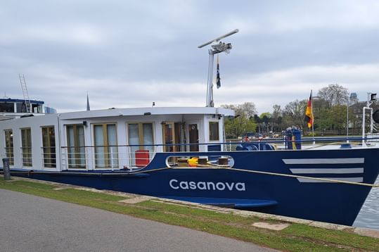 Blau-weißes Flusskreuzfahrtschiff MS Casanova am grasbewachsenen Flussufer unter bewölktem Himmel, mit Bäumen und Gebäuden im Hintergrund.
