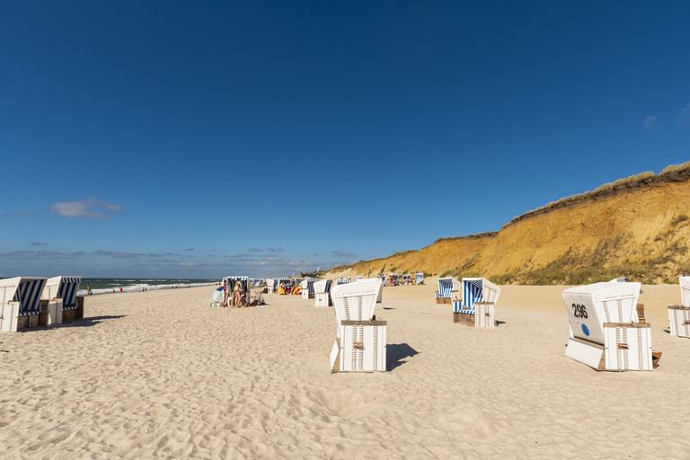 Sandstrand auf Sylt mit weiß-blau gestreiften Strandkörben verteilt im Sand. Rote Sandsteinklippe erhebt sich im Hintergrund unter klarem blauen Himmel.