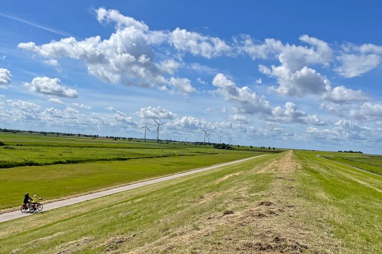 Two cyclists on a paved path along a grass-covered dike in East Frisia. Wind turbines visible in the distance under a blue sky with clouds.
