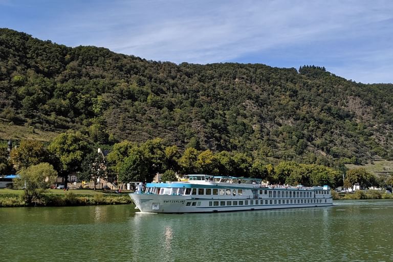 MS Switzerland Flusskreuzfahrtschiff auf ruhigem Wasser mit bewaldeten Bergen und kleinem Dorf im Hintergrund unter blauem Himmel.