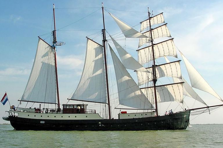 Dreimaster-Segelschiff Simmer fan Fryslan mit weißen Segeln, segelnd auf ruhigem Wasser unter blauem Himmel mit niederländischer Flagge.
