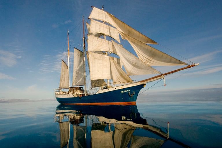 Das Segelschiff Antigua mit weißen Segeln gleitet über spiegelglattes blaues Wasser unter klarem Himmel und erzeugt eine perfekte Spiegelung.