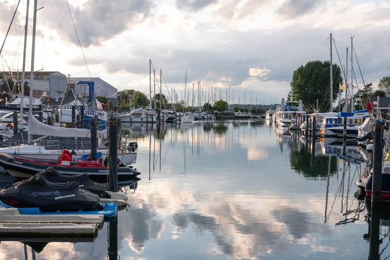 Ruhiger Yachthafen in Travemünde mit verschiedenen Booten an Holzstegen. Das ruhige Wasser spiegelt bewölkten Himmel und Masten perfekt wider.