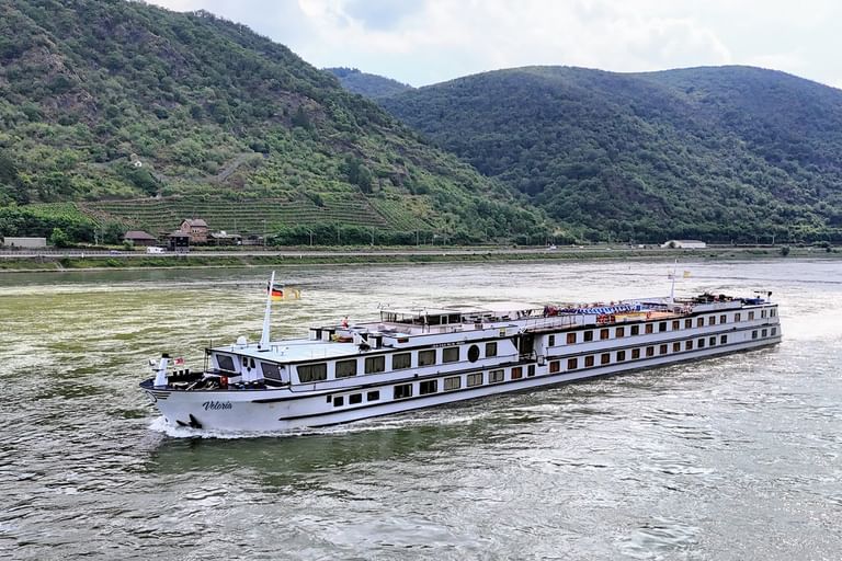 White river cruise ship MS Veloria with German flag sailing on the Rhine River, with green vineyard-covered hills in the background.