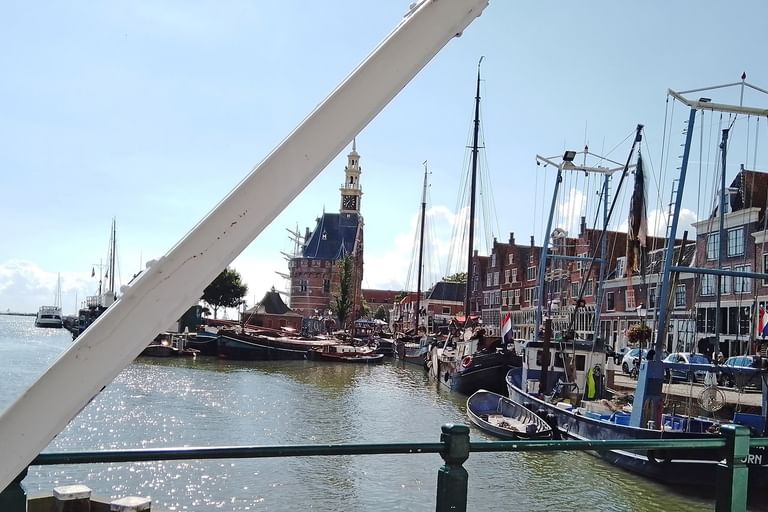 Niederländischer Hafen mit historischen Segelschiffen am Kanal. Traditionelle Gebäude und Kirchturm im Hintergrund unter blauem Himmel.