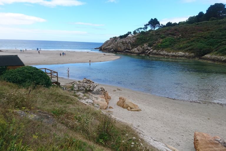 Küstenszene in der Nordbretagne mit einem Sandstrand, wo ein Fluss auf das Meer trifft, mit bewachsenen Felsklippen und spazierenden Menschen.