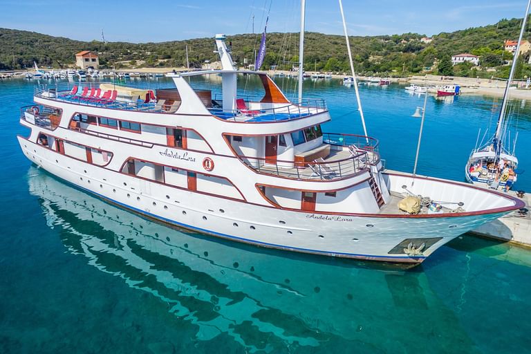 Weißes Passagierschiff MY Andela Lora mit rotem Zierstreifen am Betonsteg in türkisfarbenem Wasser. Kroatische Küstenhügel und Hafen sichtbar.