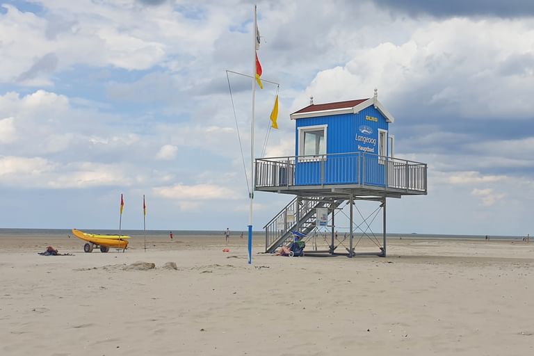 Blaue Rettungsstation am Strand von Langeoog Blaue erhöhte Rettungsstation auf Stelzen am Strand von Langeoog mit deutschen Flaggen, Treppe und gelbem Boot in der Nähe auf sandigem Ufer.