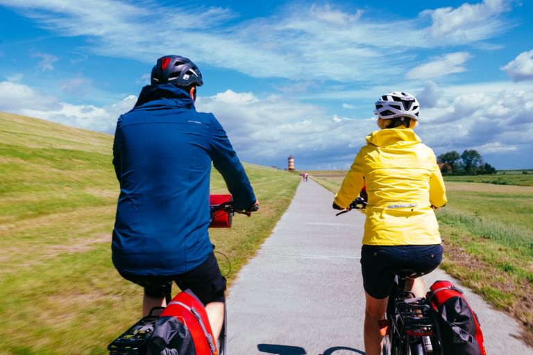 Zwei Radfahrer mit Packtaschen fahren auf gepflastertem Deichweg in Ostfriesland. Einer trägt blaue, der andere gelbe Jacke. Rot-weißer Leuchtturm Pilsum voraus sichtbar.