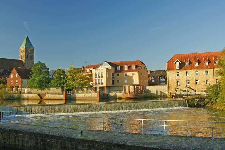 Flussblick auf Rheine mit Wehr, historischen Gebäuden einschließlich Kirchturm, bunten Häusern entlang der Uferpromenade unter blauem Himmel.