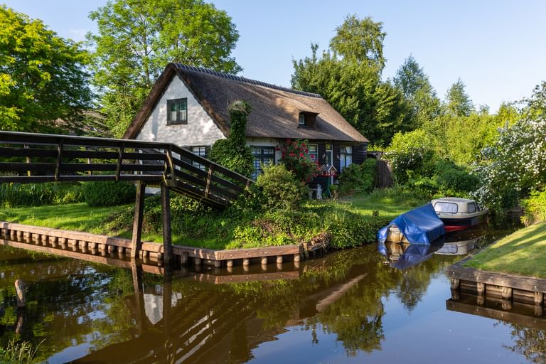 Traditionelles Reetdachhaus neben einem Kanal in Giethoorn mit Holzbrücke und Booten am Ufer, umgeben von üppigem Grün.