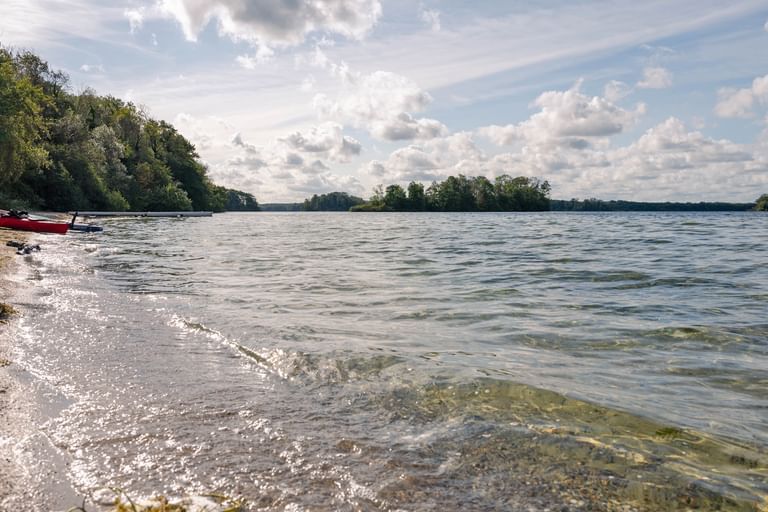 Sandstrand in Travemünde mit sanften Wellen am Ufer. Rote und weiße Boote liegen am Strand, dichter Wald säumt das Ufer.