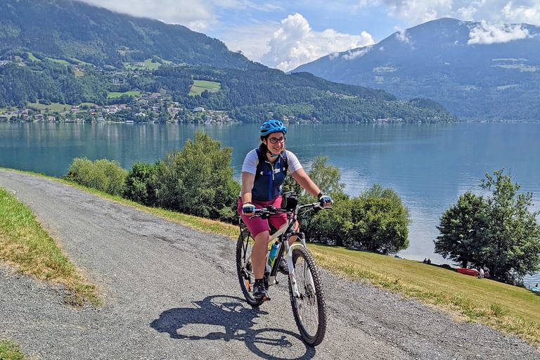 Radfahrerin mit blauem Helm fährt auf befestigtem Weg entlang großem Alpensee mit bewaldeten Bergen und Dörfern im Hintergrund.