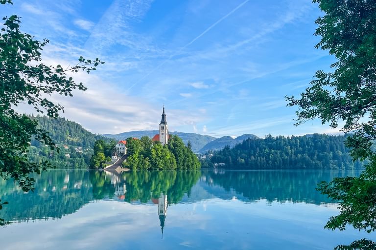 Bleder See in Slowenien mit kleiner Insel mit Kirche und weißem Glockenturm, umgeben von bewaldeten Hügeln und Bergen unter blauem Himmel.