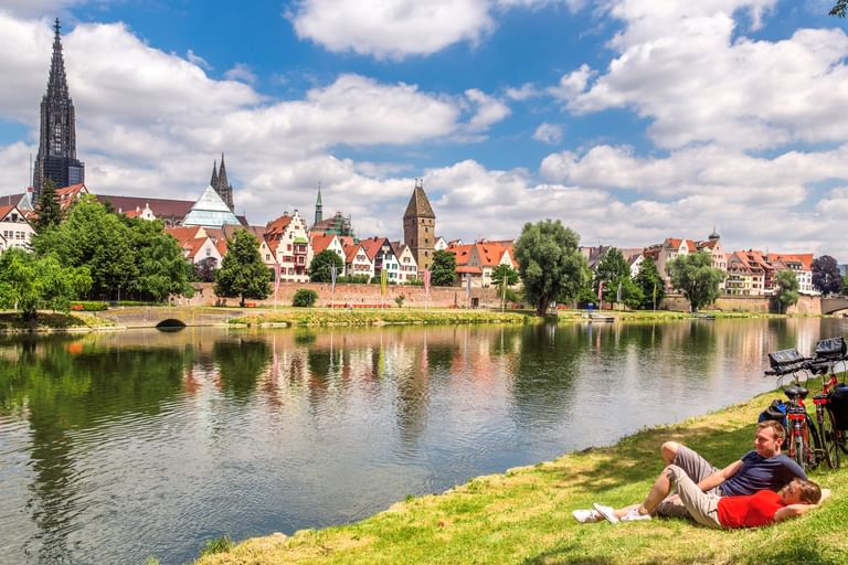 Zwei Radfahrer ruhen im Gras an der Donau in Ulm, mit dem gotischen Ulmer Münster und historischen Gebäuden im Wasser gespiegelt.
