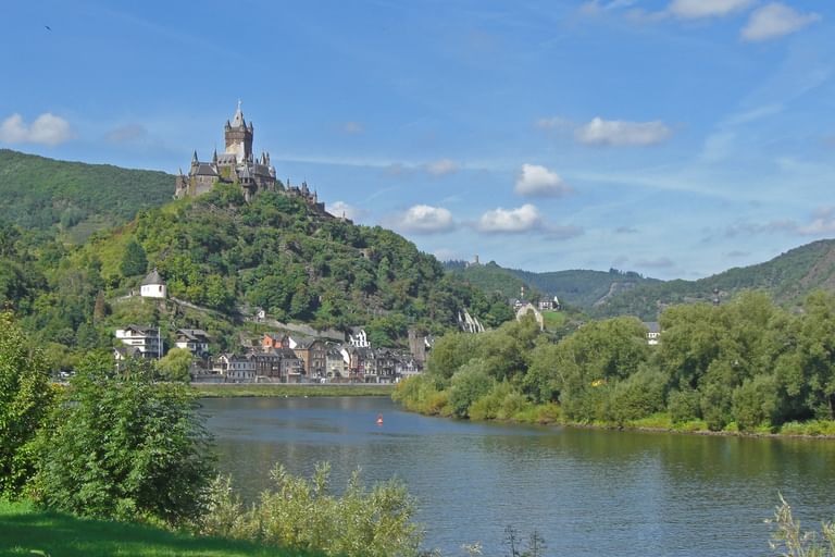 Burg Cochem thront auf einem bewaldeten Hügel über der Mosel, mit der Stadt Cochem am Flussufer darunter.