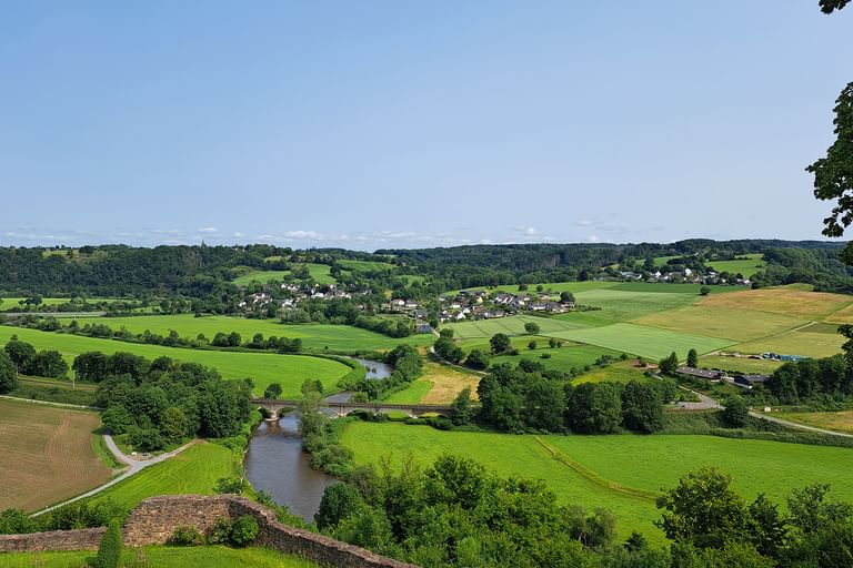 Panoramablick auf einen gewundenen Fluss durch grüne Felder und Ackerland bei Bonn. Ein kleines Dorf liegt im Tal mit Wäldern auf sanften Hügeln.