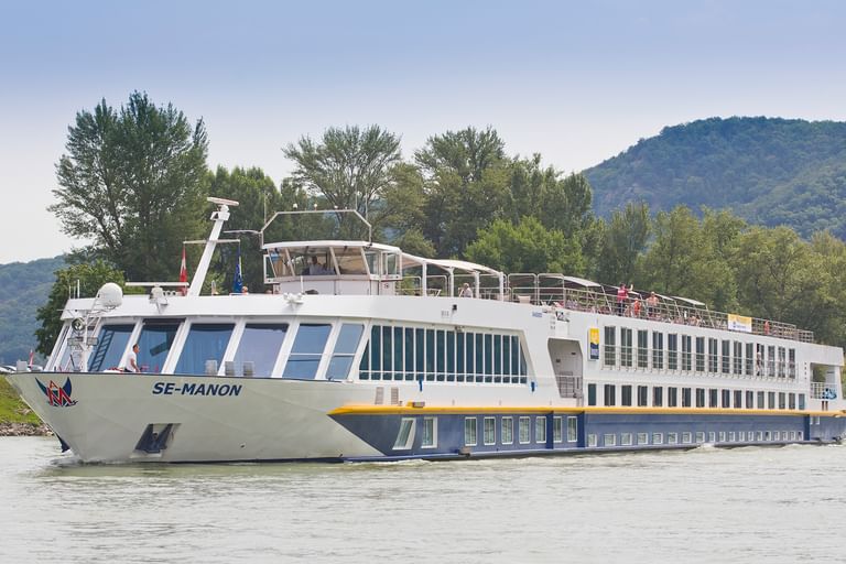 Weißes Flusskreuzfahrtschiff SE-Manon fährt auf der Donau mit Passagieren an Deck, bewaldete Hügel und grünes Flussufer im Hintergrund.
