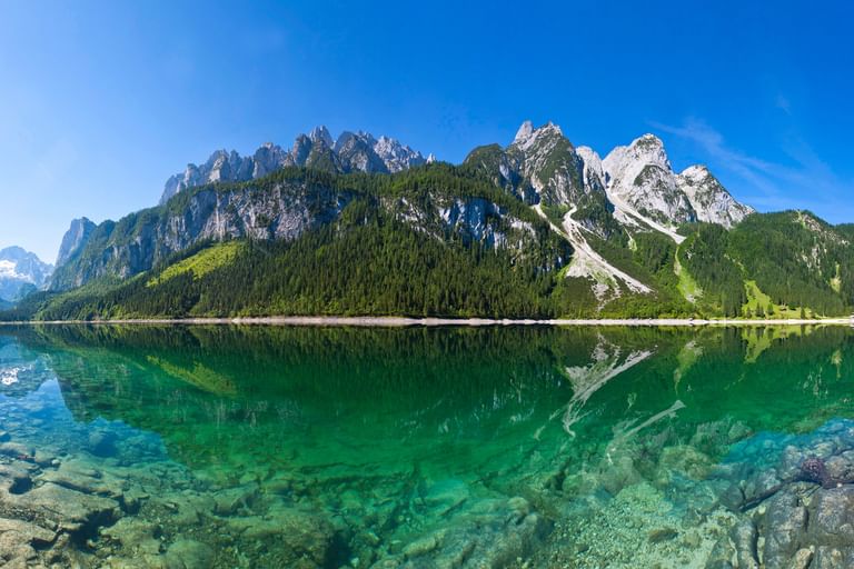 Kristallklarer Gosausee im Salzkammergut mit smaragdgrünem Wasser, das dramatische Alpengipfel unter blauem Himmel spiegelt.
