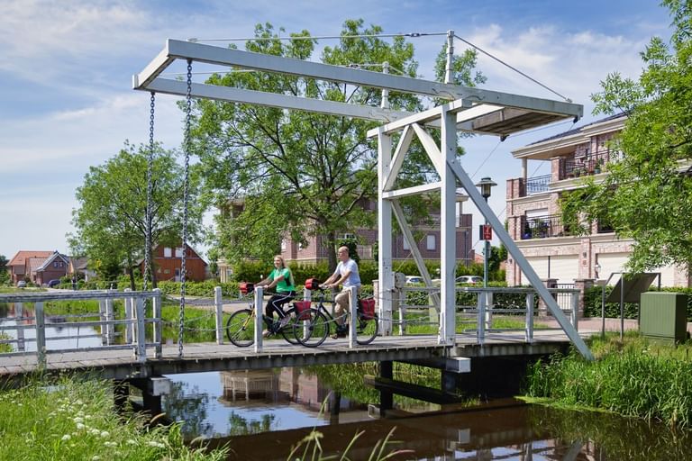Zwei Radfahrer mit Rädern stehen auf einer weißen Metall-Zugbrücke über einem Kanal. Traditionelle holländische Häuser und grüne Bäume im Hintergrund.
