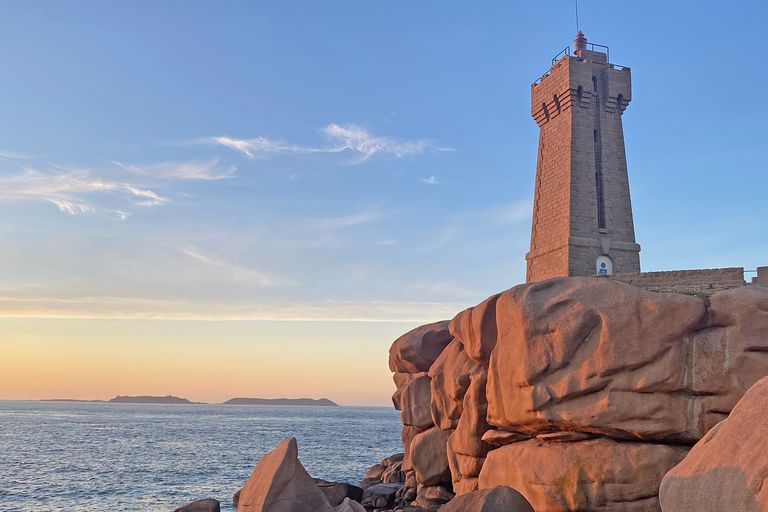 Steinerner Leuchtturm auf rosa Granitfelsen an der Küste bei Sonnenuntergang. Das Meer und entfernte Inseln sind unter blauem Himmel mit Wolken sichtbar.