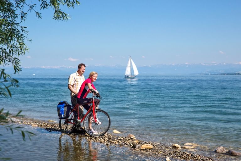 Zwei Radfahrer mit rotem Fahrrad stehen am steinigen Ufer des Bodensees. Weißes Segelboot auf blauem Wasser mit Bergen im Hintergrund.