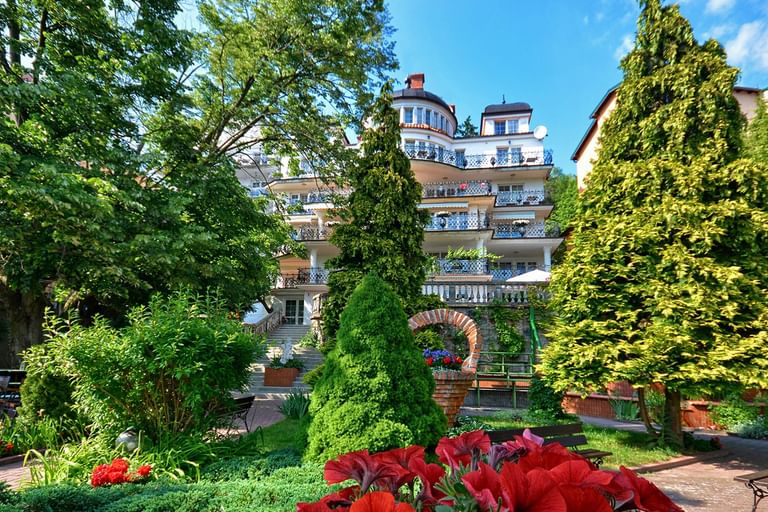 Mehrstöckiges Hotel in Masuren mit Balkonen, umgeben von üppigem Garten mit hohen Bäumen, geformten Koniferen und roten Blumen im Vordergrund.